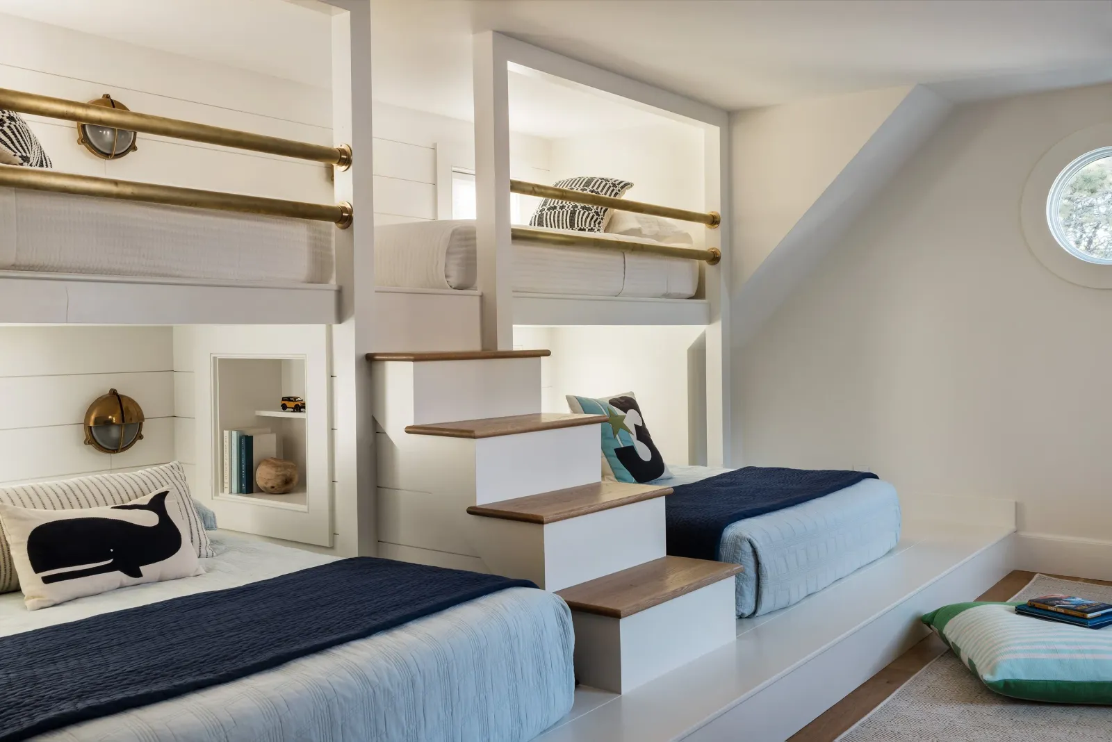 Two beds with navy blue covers and whale-themed pillows in a white, elevated bedroom with wood stairs, built-in book shelves, and a circular window.