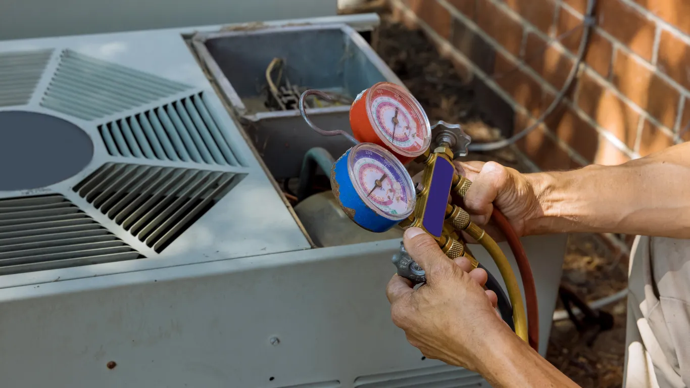 A person is using a manifold with gauges and hoses to service an HVAC unit outside. The person's hands are visible, and the unit is situated next to a brick wall.