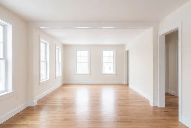 Home remodeling: long empty white room with plain cased openings, side windows and oak flooring