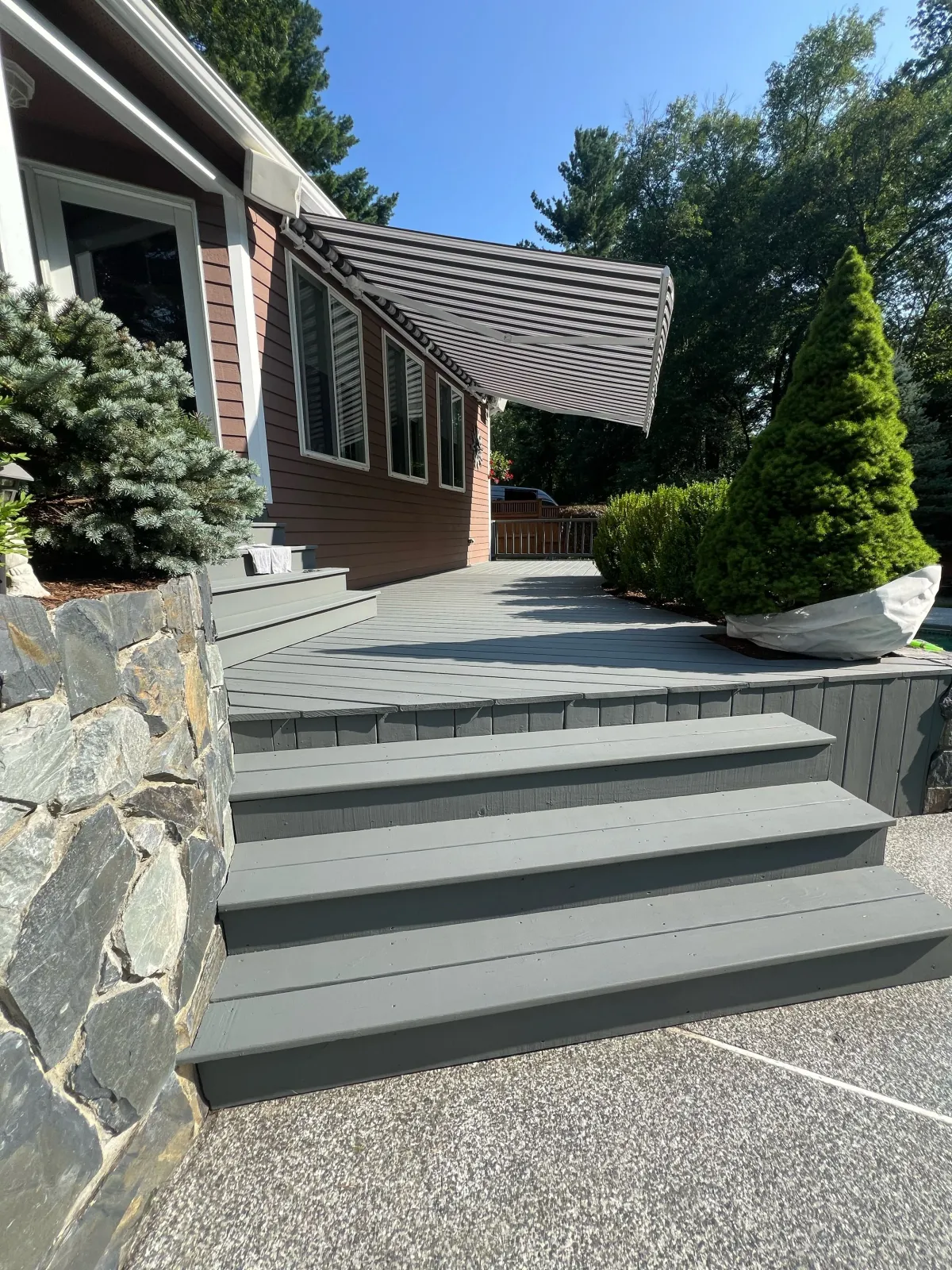 Gray wooden deck and stairs leading to the back door of a house with a striped awning, surrounded by green bushes and trees.