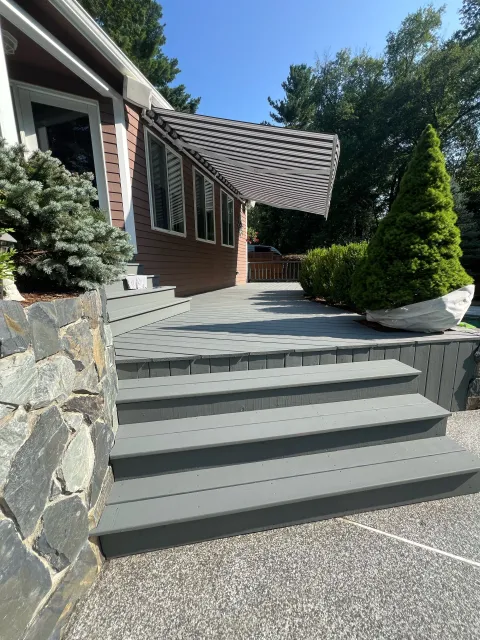 Gray wooden deck and stairs leading to the back door of a house with a striped awning, surrounded by green bushes and trees.