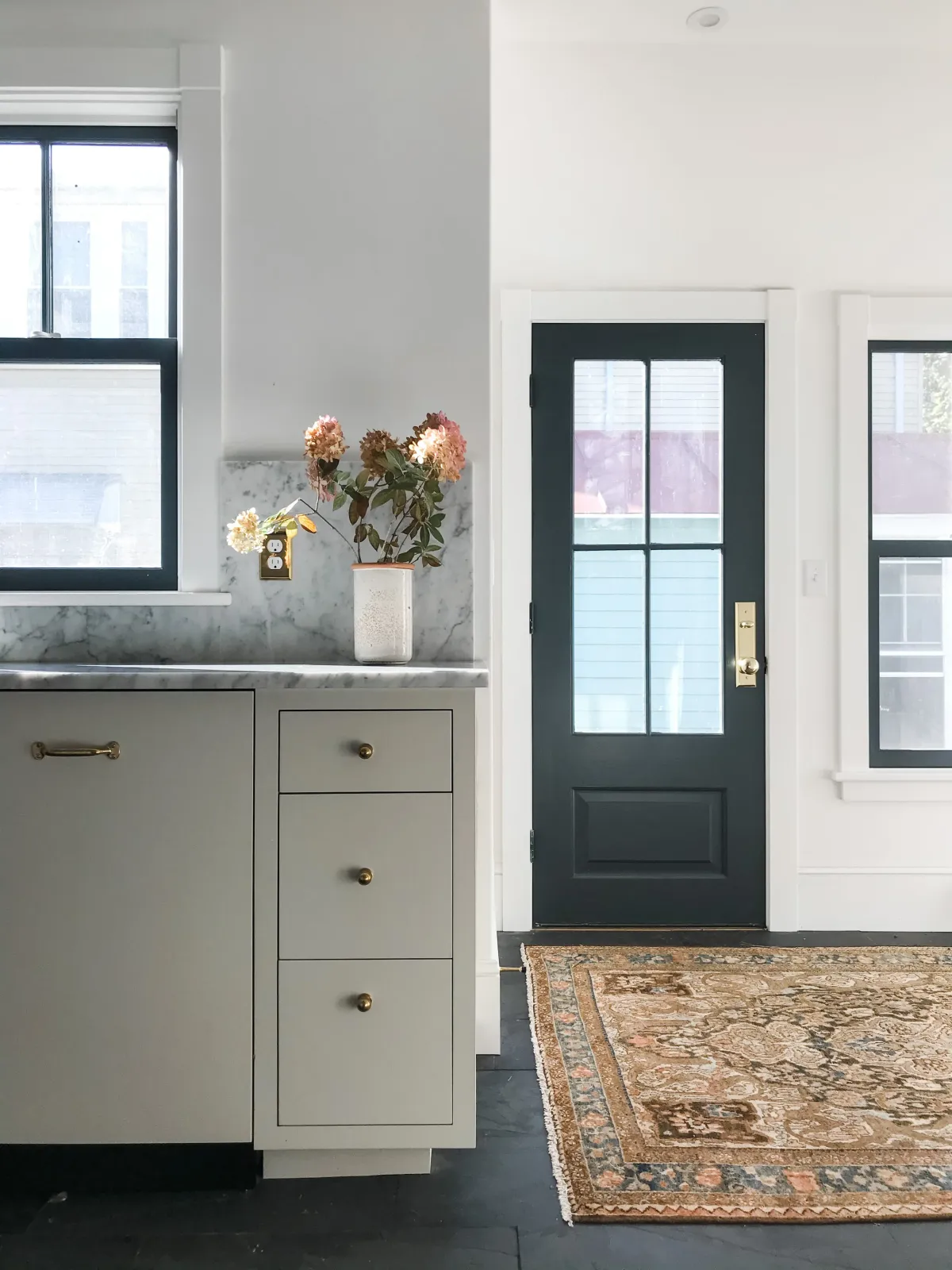 Interior view of a house entryway showing a window, a dark door with a glass window pane, a rug on the floor, and a cabinet with a potted flower on top.