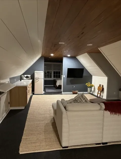 Cozy attic living room with sloped ceilings, sofa, TV, rug, and small kitchenette area.