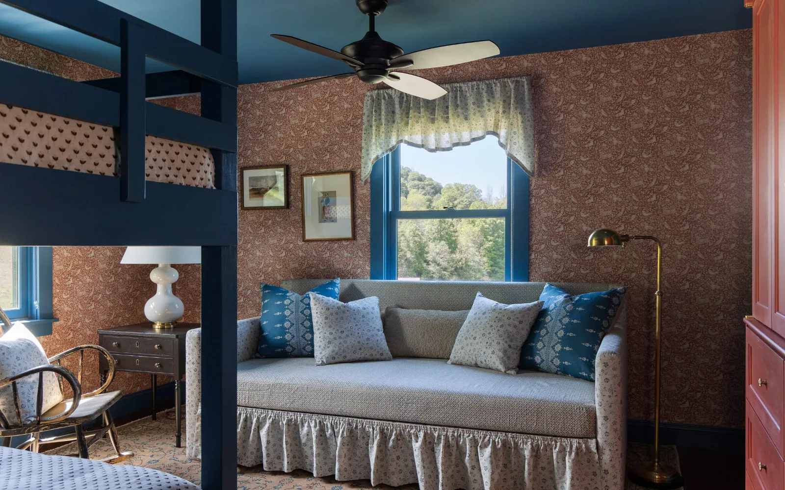 A cozy seating nook with a ruffled daybed and blue pillows, located next to a window overlooking trees, framed by brown patterned wallpaper and blue trim.