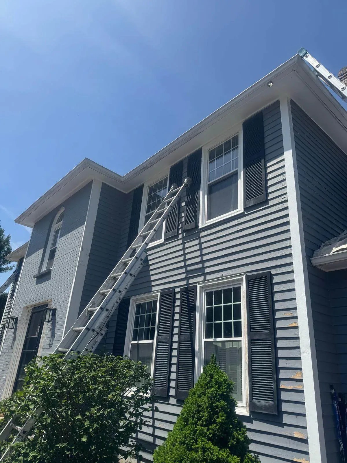 Tall gray house with white trim and black shutters, where a long ladder leans against the wall reaching the second-floor windows. Paint patches and tools suggest ongoing exterior painting or repair work under clear blue skies.