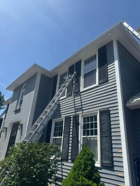 Tall gray house with white trim and black shutters, where a long ladder leans against the wall reaching the second-floor windows. Paint patches and tools suggest ongoing exterior painting or repair work under clear blue skies.