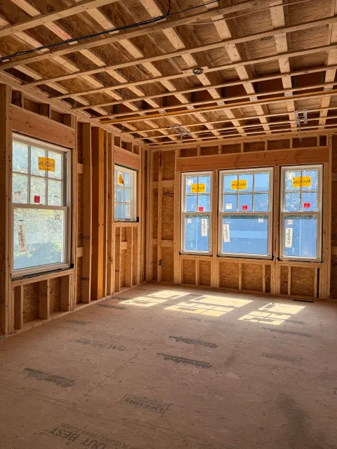 Newly constructed room with exposed wooden framing, three double-pane windows, and sunlight casting shadows on the unfinished floor.