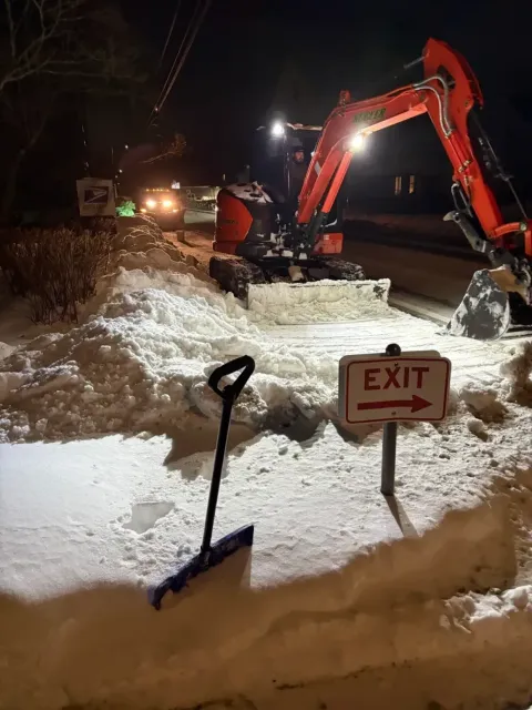 Snowplow clearing snow at night beside an EXIT sign, with a shovel in the foreground.