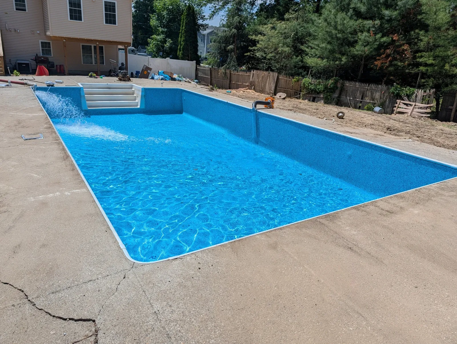 A rectangular swimming pool with blue interior and white steps under construction in a backyard, surrounded by unfinished concrete surface and debris, with a house and trees in the background.
