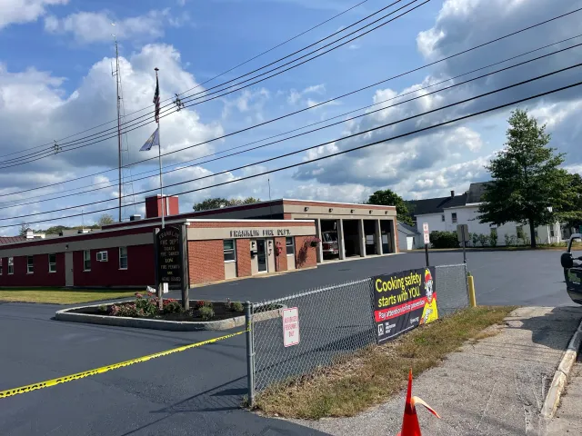 Front view of Franklin Volunteer Fire Department with a parking lot, a chain-link fence, and a banner that reads 'Cooking safety starts with you. Pay attention to fire prevention.' There are power lines in the sky, a flagpole with the American flag and another flag, a small planting bed with flowers, and a single traffic cone.