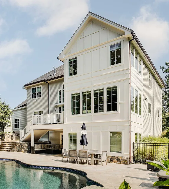 Three-story white home exterior with black-framed windows, second-floor covered deck, and ground-level patio with umbrella seating.