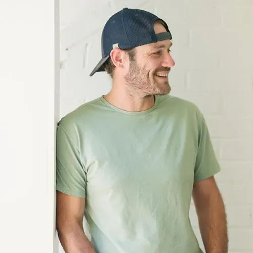 Smiling man in a backward navy cap and light green T-shirt standing indoors by a white wall
