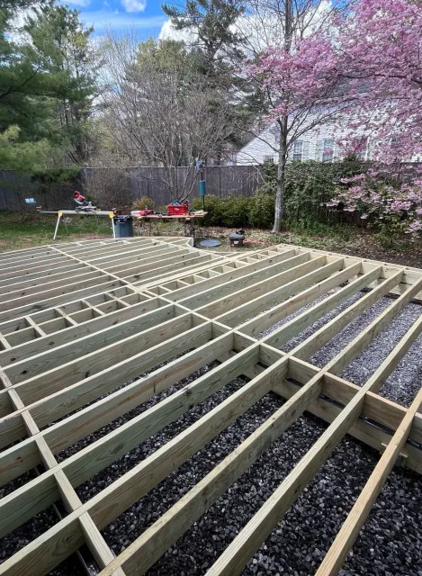 Wooden deck framing in a backyard with a blooming pink tree, a fenced yard, and tools on work tables.