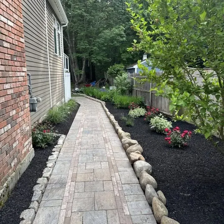 Stone walkway bordered by rocks and garden beds with flowers and greenery beside a house and wooden fence.