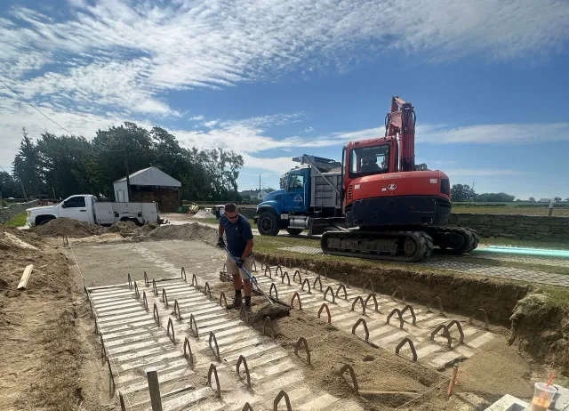 A person works on a construction site with rows of steel rebar protruding from concrete footings, an excavator, and trucks.