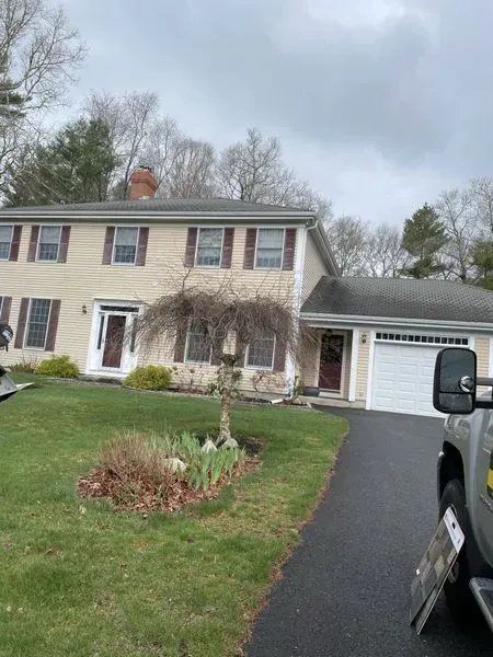 Two-story beige house with brown shutters and a weeping tree in front, on a green lawn with a black driveway.