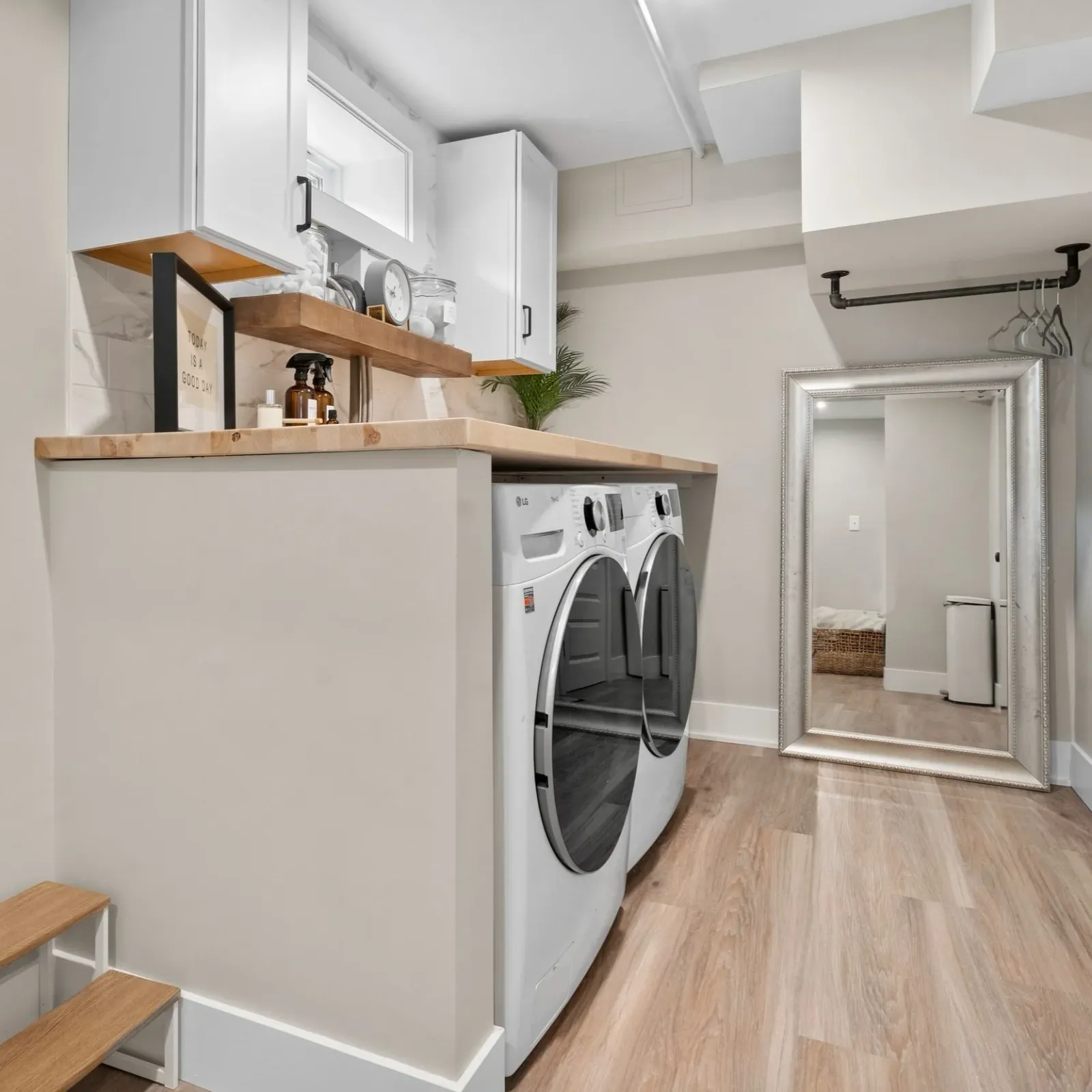 A laundry room with white washer and dryer, a wooden countertop, a large mirror leaning against the wall, and a small step stool.