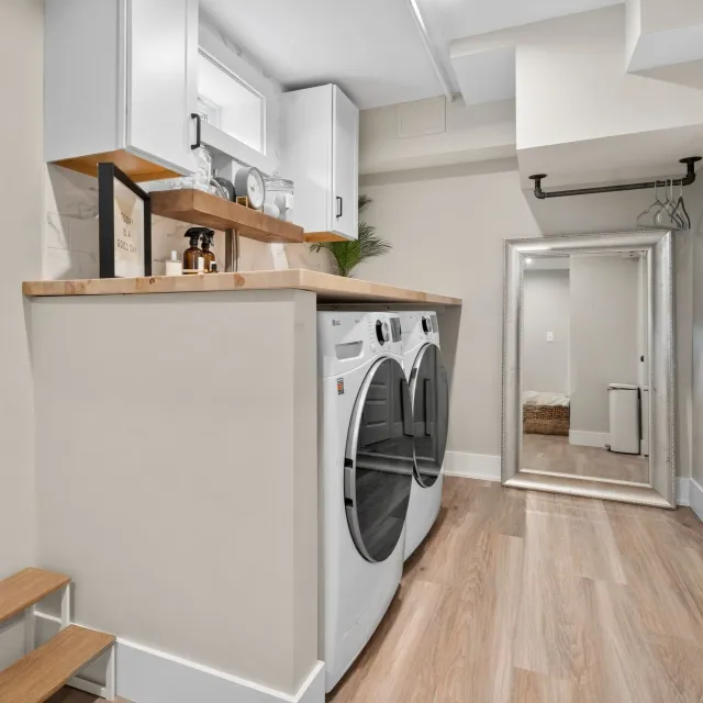 A laundry room with white washer and dryer, a wooden countertop, a large mirror leaning against the wall, and a small step stool.