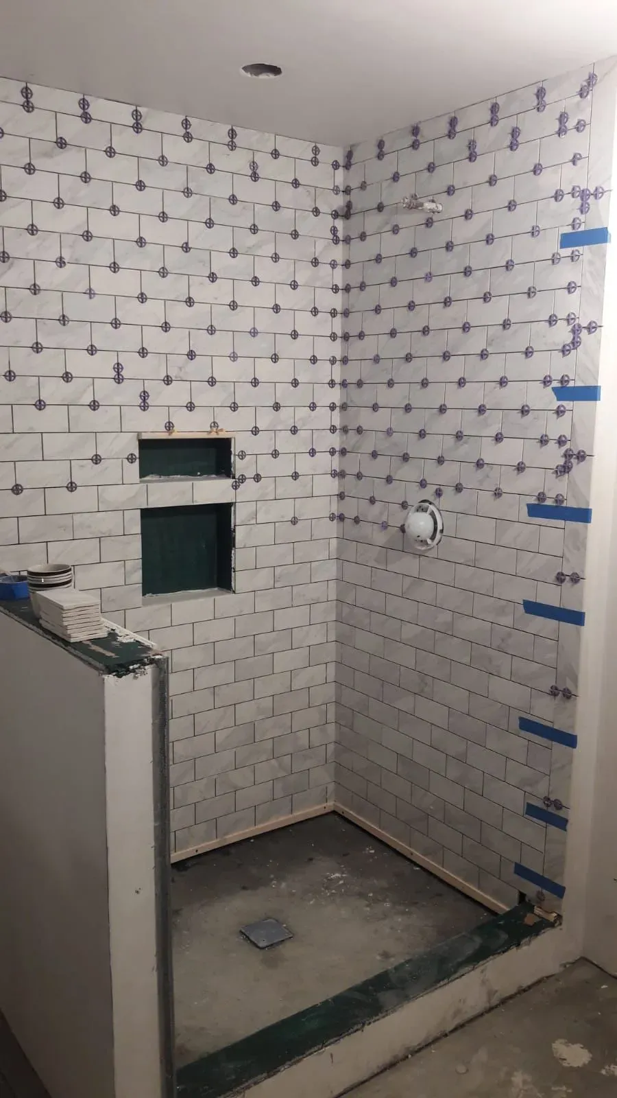 Shower tile installation progress showing marble look subway tiles and leveling spacers around two wall niches.