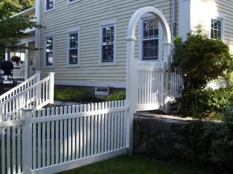 A white picket fence and a matching arbor gate sit in a grassy yard beside a yellow house with stone foundation walls.