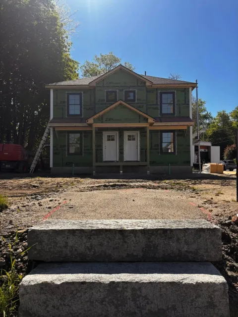 Green two-story house under construction with gravel front yard and concrete steps in sunlight