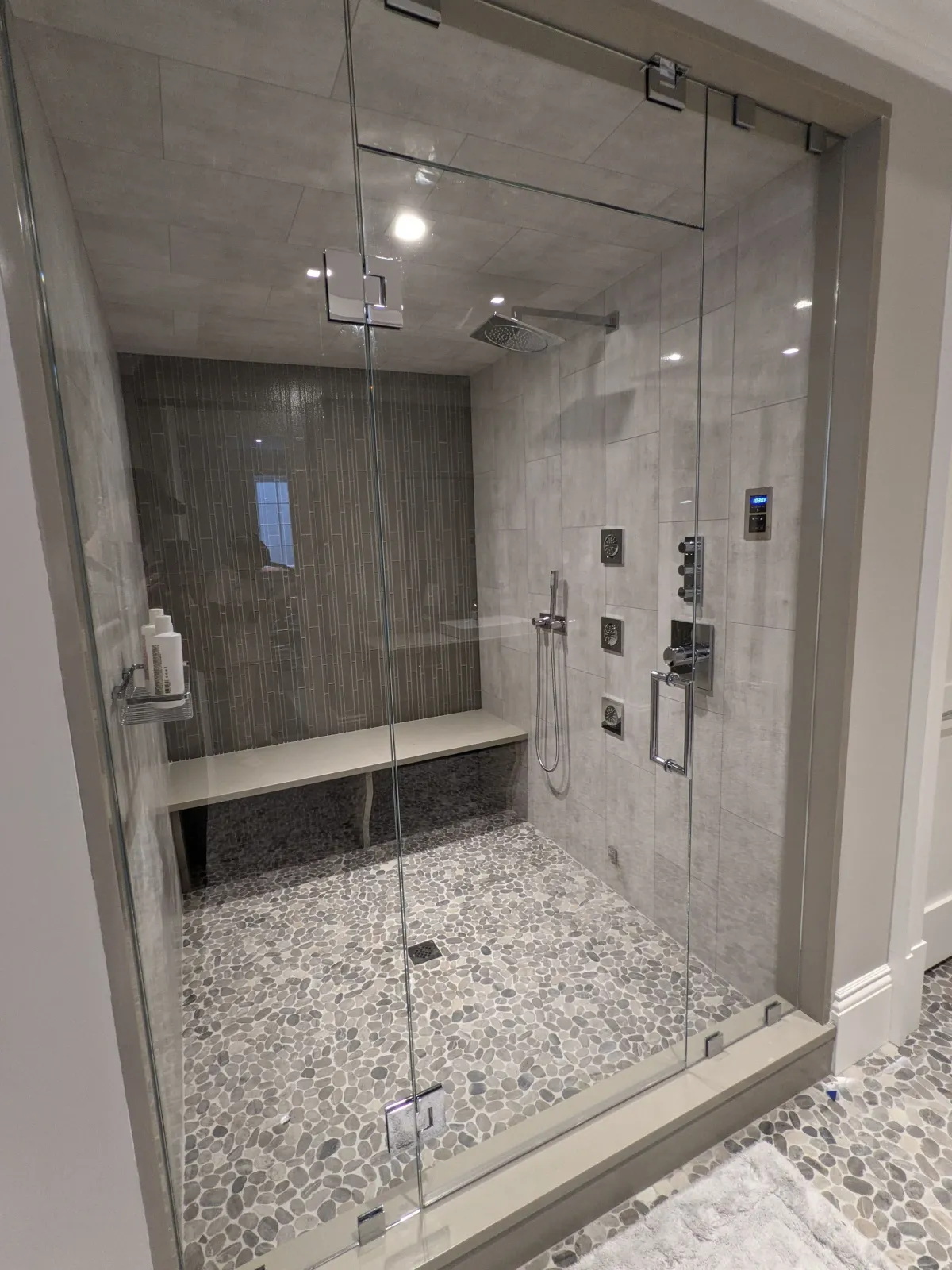 Modern walk-in shower with glass enclosure, pebble floor, built-in bench, and multiple shower heads and controls.