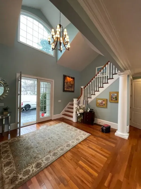 Bright foyer with chandelier, staircase, hardwood floors, large rug, and glass front door.