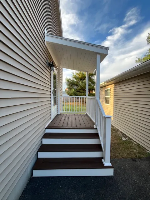 A small porch with wooden stairs leading up to a door, with white railings and beige siding house walls, under a covered roof, in a suburban backyard with trees and a partly cloudy sky.