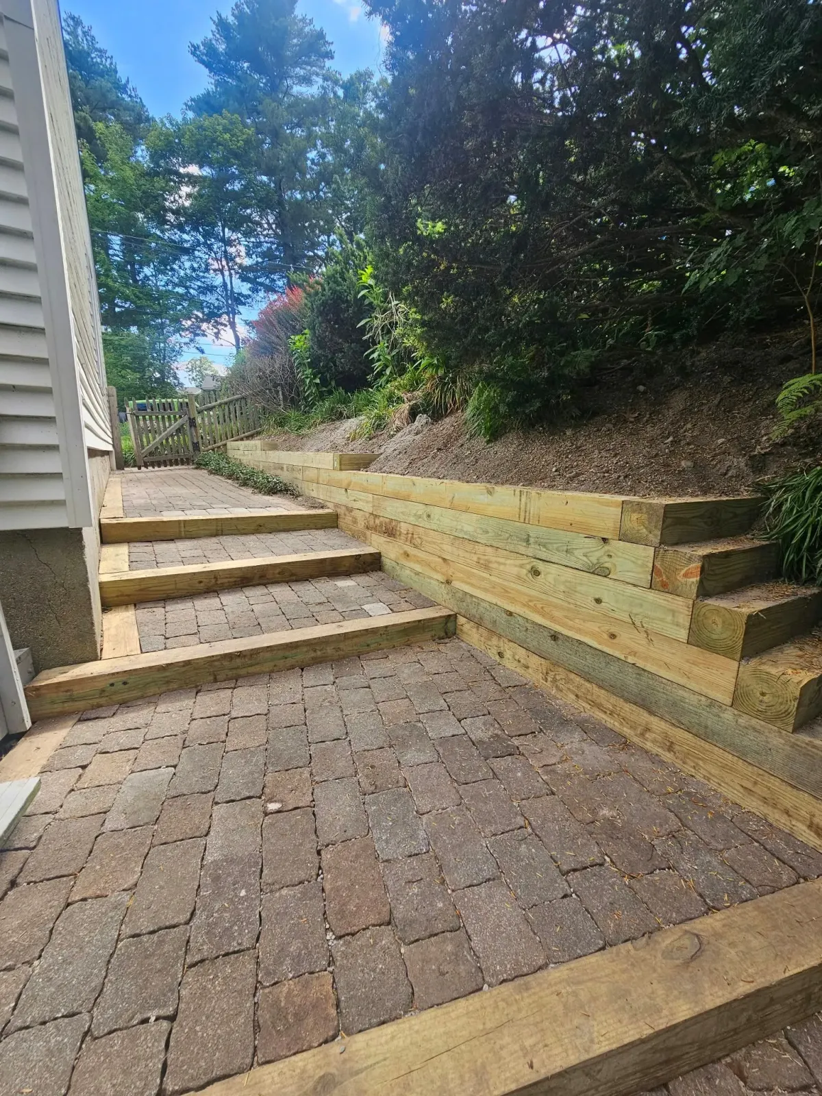 Brick patio with steps beside a house and retaining wall, lined by trees and shrubs under a blue sky