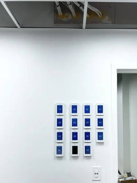 A wall with several thermostat panels on for controlling multiple floors and offices