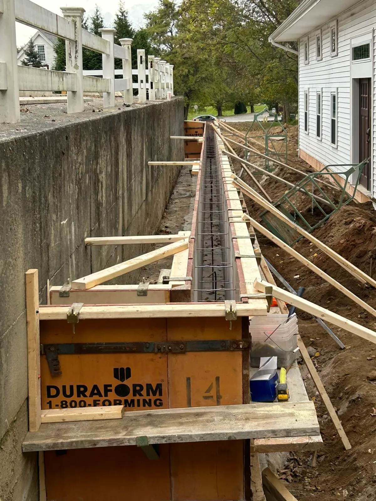 Construction site with scaffolding on a sidewalk, work spanning a retaining wall with stakes and wood beams, next to a white house and a fenced yard, with trees and a road in the background.