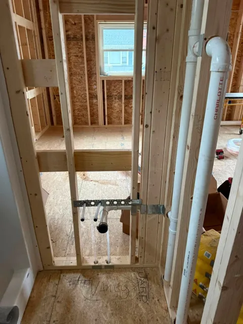 Interior of a house under construction, with exposed wall framing and plumbing pipes near a hallway doorway.