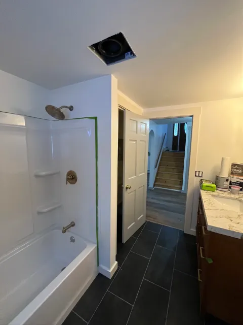 Interior view of bathroom, showing a shower with white walls, a brass showerhead, and black floor tiles. In the background, stairs lead to an upper level, and a cabinet with painting supplies and a lamp are on the right.