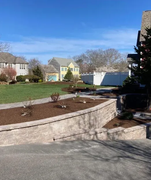 Stone retaining wall with mulch and young plants, leading to a lawn with houses in the background.
