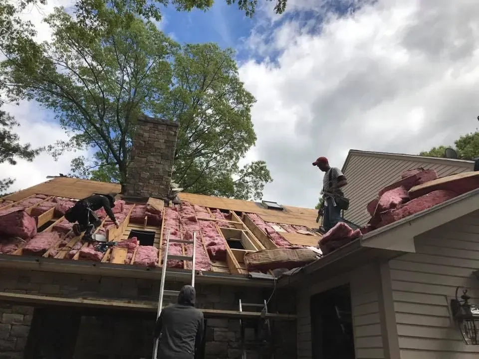 Roofers repairing a house roof, removing old shingles and insulation. Chimney visible. Cloudy sky.