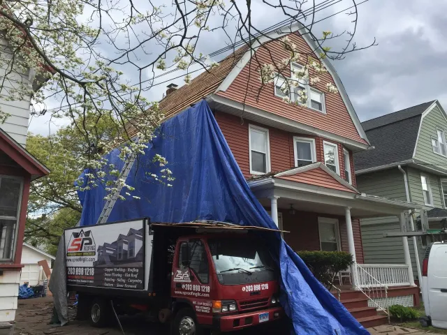 A red roofing truck parked in front of a brick house, with a blue tarp draped over the roof to cover repair work.