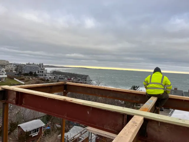 A construction worker in a bright yellow safety jacket and dark hood sits on a steel beam frame of a building under construction, overlooking a coastal neighborhood with houses and the ocean in the distance during a cloudy day.