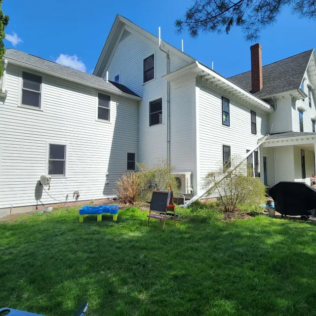 White two-story house with gray roof, green lawn, and a small patio chair under a blue sky.