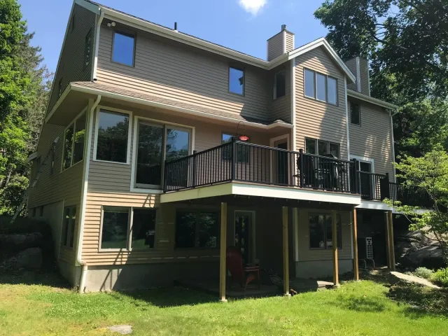 Two-story tan home with new exterior paint by Star Painting, featuring a black balcony and large windows in a wooded New England setting