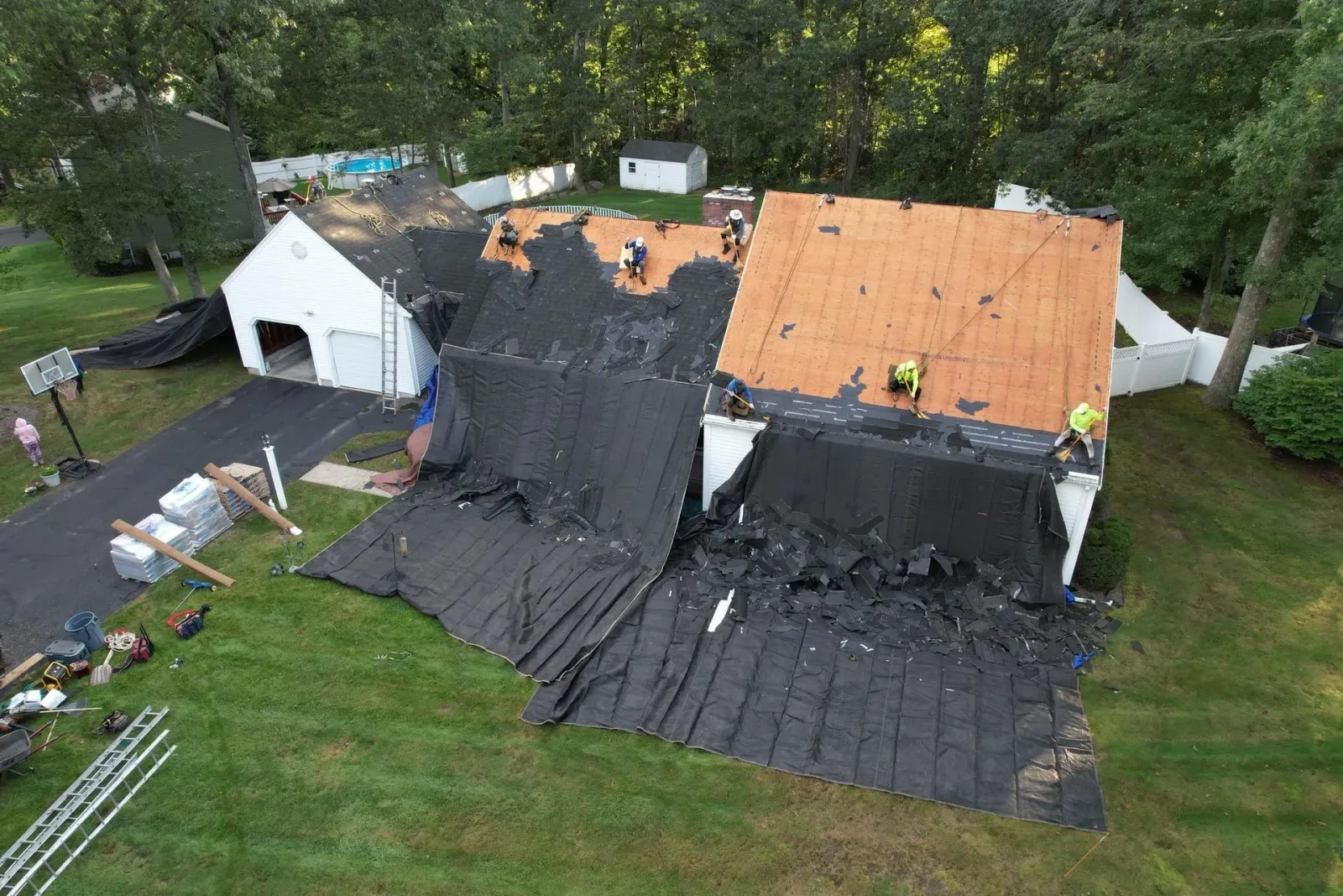 Damaged roof covered with black tarp after storm, with orange plywood patches on a house in a grassy yard