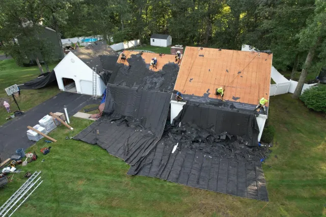 Damaged roof covered with black tarp after storm, with orange plywood patches on a house in a grassy yard