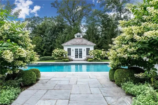 A backyard with a swimming pool, surrounded by lush green bushes and trees, and a small white pool house in the background.