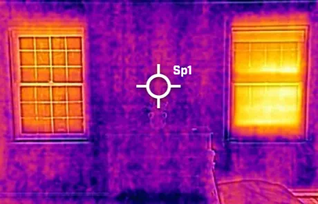 Infrared thermal image of a house wall with two windows emitting heat; the wall and frames appear in purple and yellow indicating temperature differences.