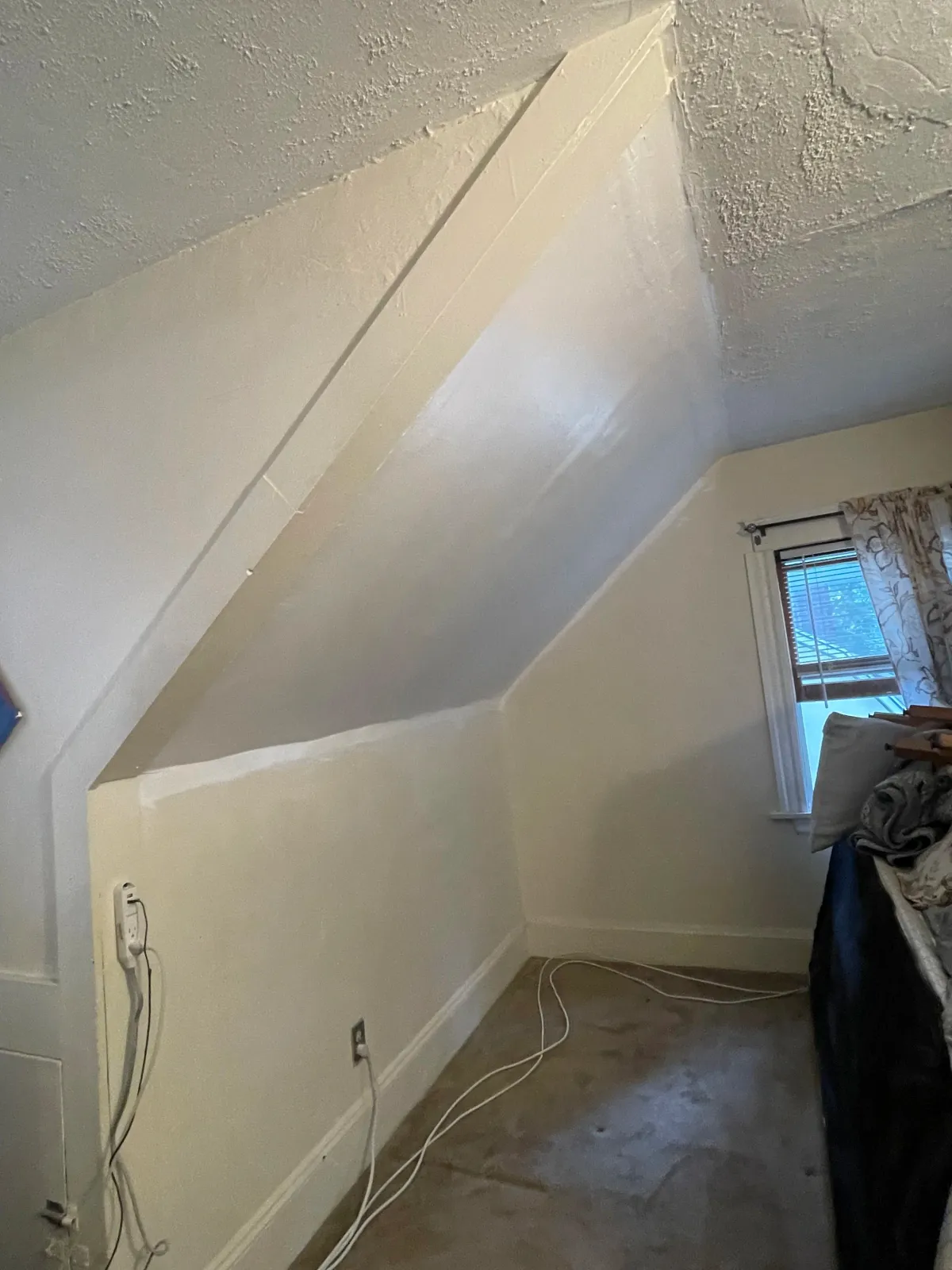 A room with a sloped ceiling and window with floral curtains, wires and cords hanging loosely near wall outlets, and a pile of clothes on a piece of furniture.