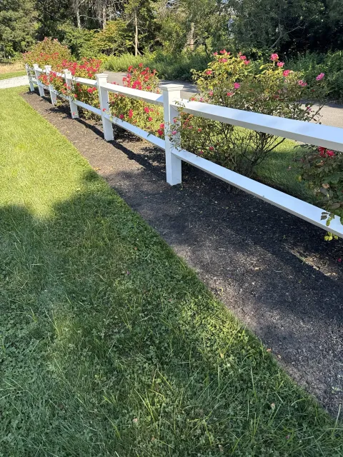 White wooden fence along a sidewalk with pink flowering bushes, green grass, and trees in the background.