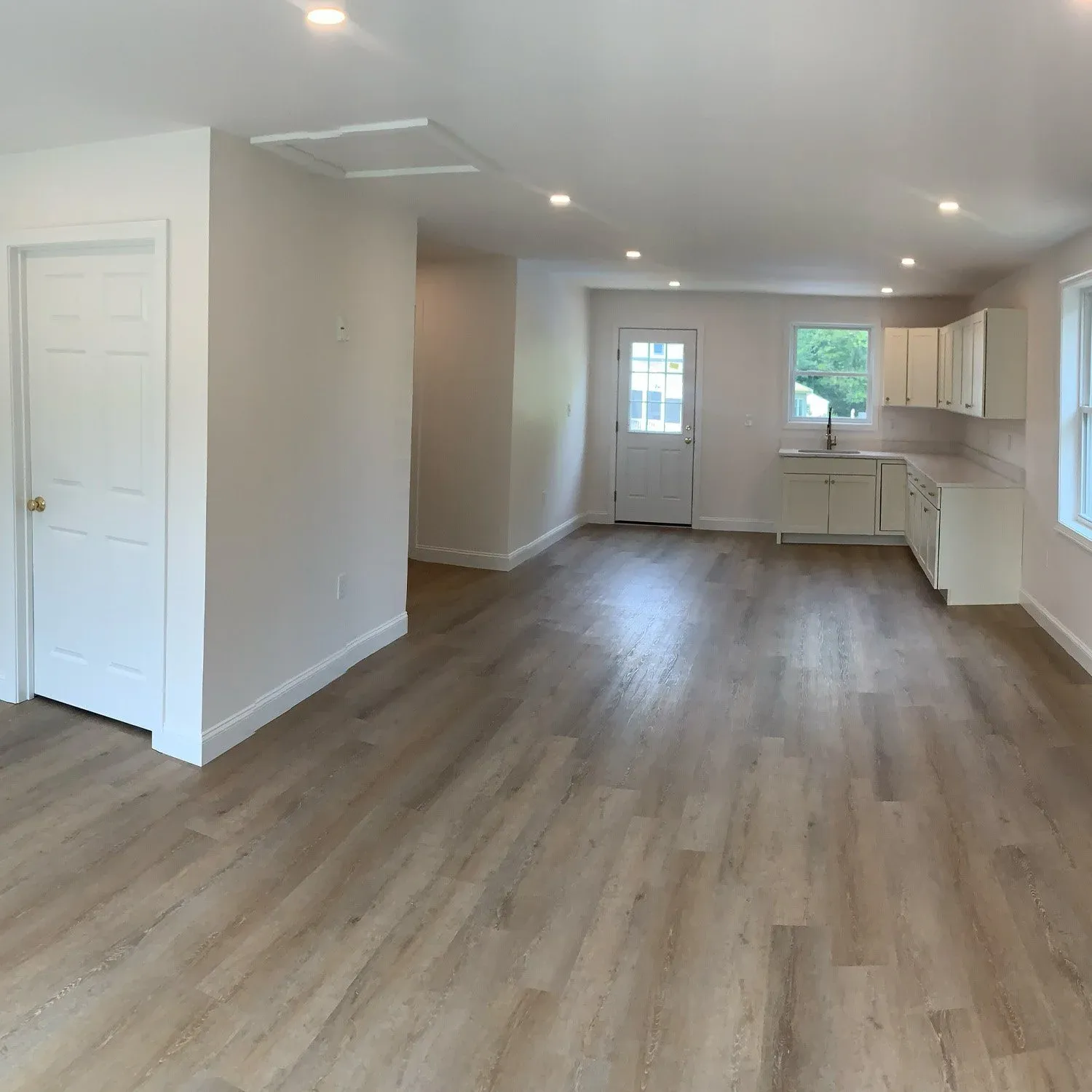 Empty living room and kitchen area with hardwood floors, white walls, and multiple windows letting in natural light.