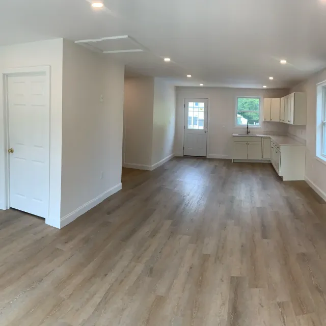 Empty living room and kitchen area with hardwood floors, white walls, and multiple windows letting in natural light.