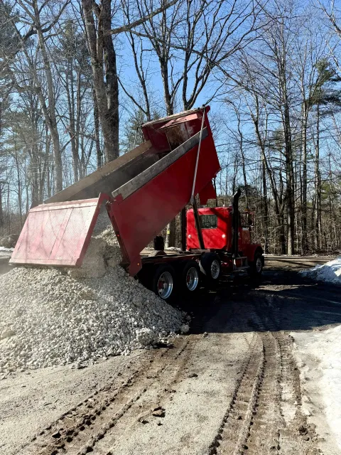 Red dump truck tipping gravel onto a dirt road in a wooded area