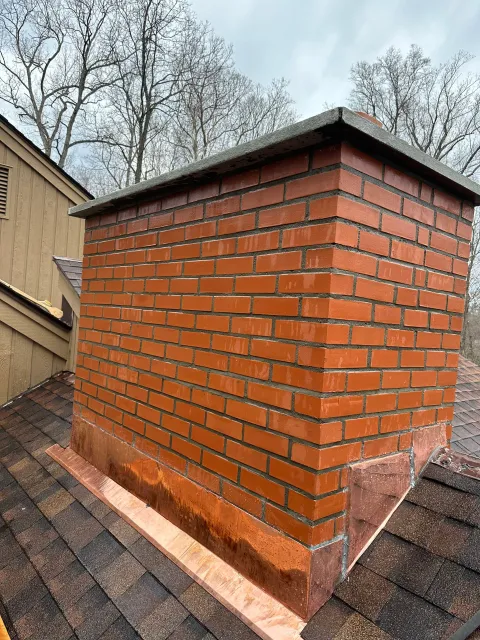A brick chimney on a shingled roof with new copper flashing installed at the base.