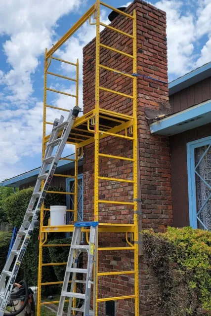 Yellow scaffold beside a brick chimney under repair, with ladders and a bucket outside a house